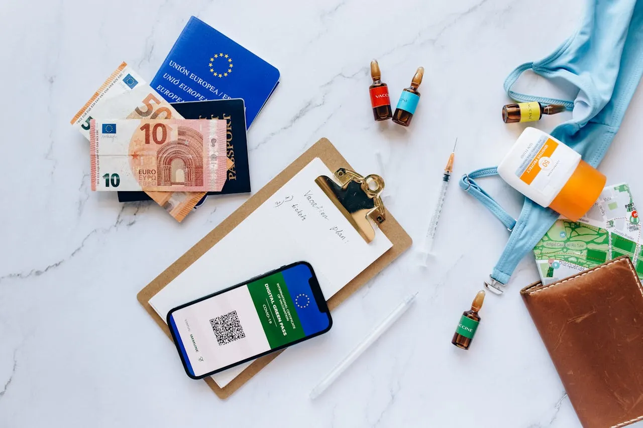 Flat-lay of trip paperwork on a marble surface: a passport, euro notes, a handwritten plan on a clipboard, a phone showing a digital health certificate, vaccine ampoules and a syringe, a folded map, sunscreen and a leather wallet