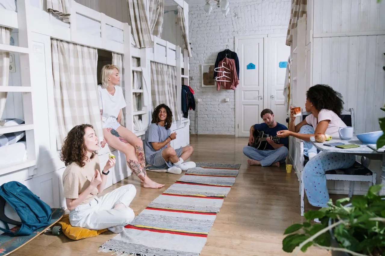 Travelers sitting on bunks and the floor of a bright hostel dorm, chatting while one of them plays guitar