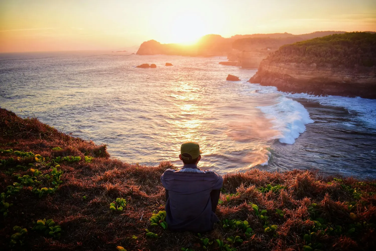 A person in a cap sitting on a grassy clifftop at sunset, looking out over a curving coastline with waves breaking below